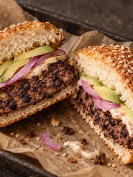 A homemade smoky bean burger cut in half, with avocado, pickled red onion and lime mayonnaise in a sesame bun.